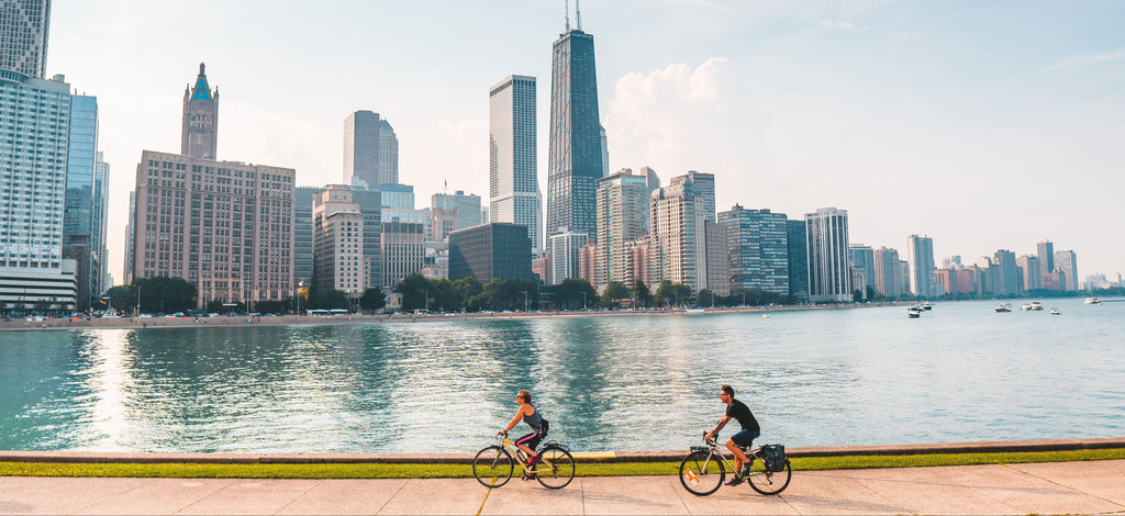 Two cyclists ride along a waterfront promenade with a large city skyline across the water under a pale blue sky.